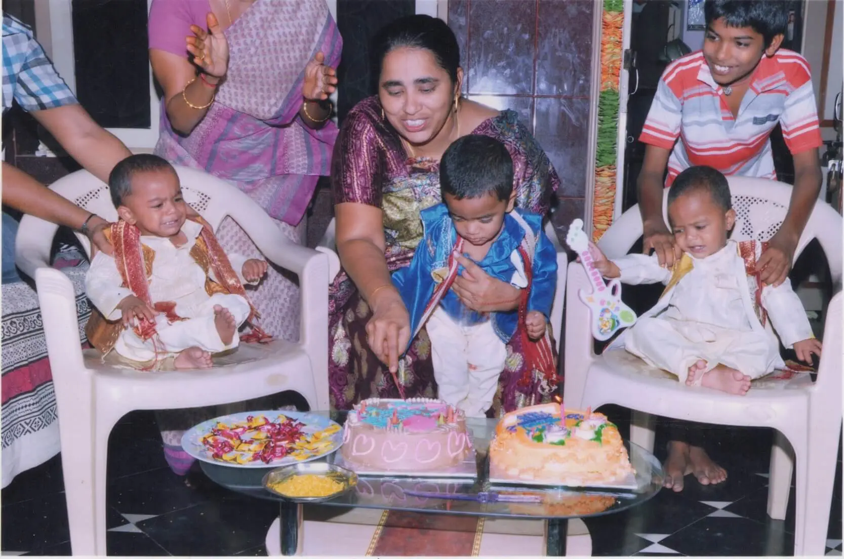 Three infants, born through ICSI at Karthika Datta's IVF clinic in 2010, seen celebrating their first birthday. Mother seated in a chair and holding one son and making him cut the cake, flanked by the other two children sitting in chairs on each side.