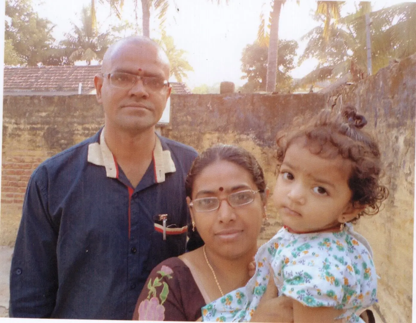 Father (to the left), mother (in the middle), holding a young girl born through ICSI at Karthika Datta's IVF clinic, against a backdrop of tiled roofs and coconut trees.