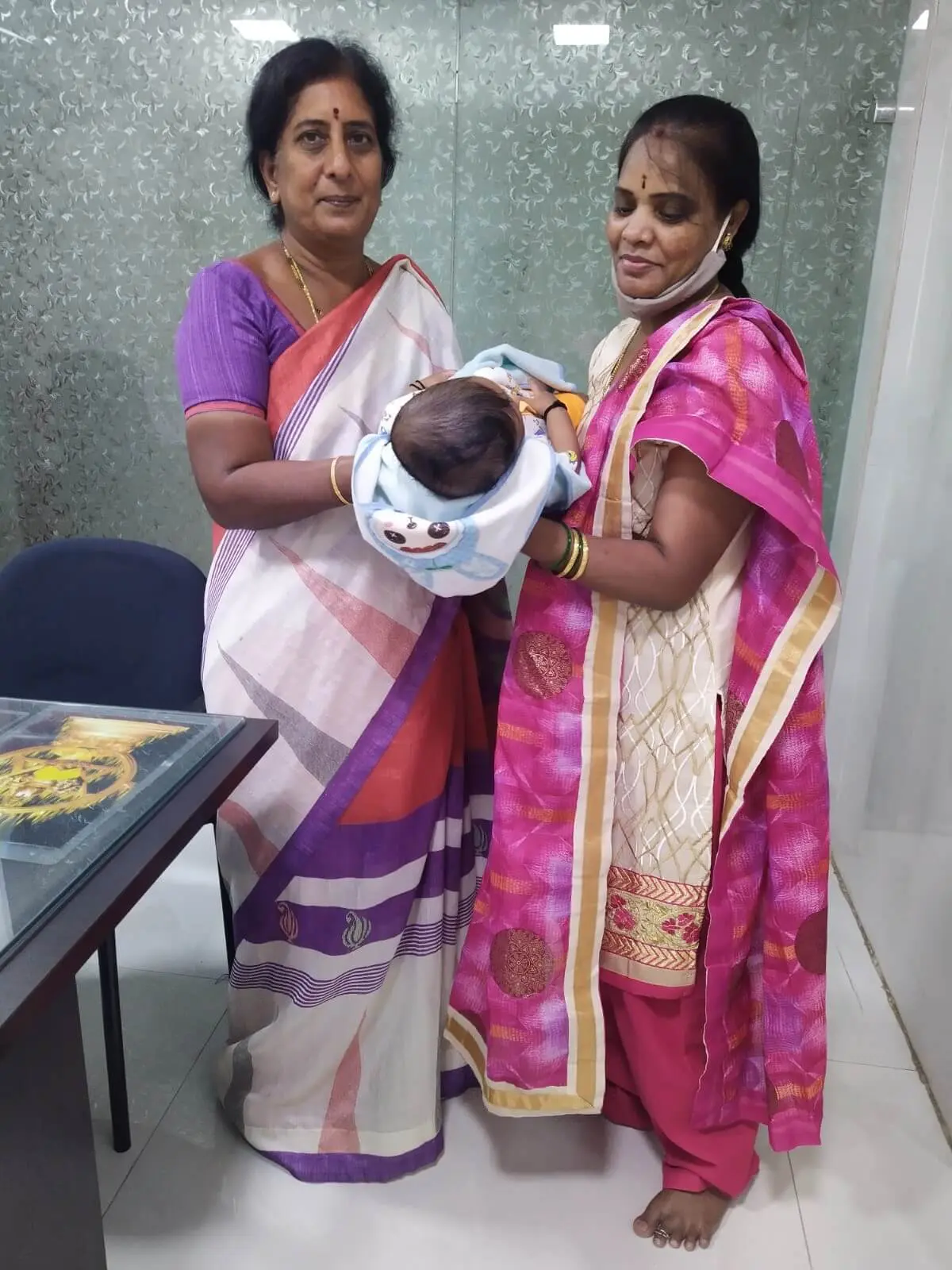Dr. Rama Devi Kolli (L), standing and holding an infant, along with the mother (R), against the backdrop of glass doors, in a consultation room at Karthika Datta's IVF.